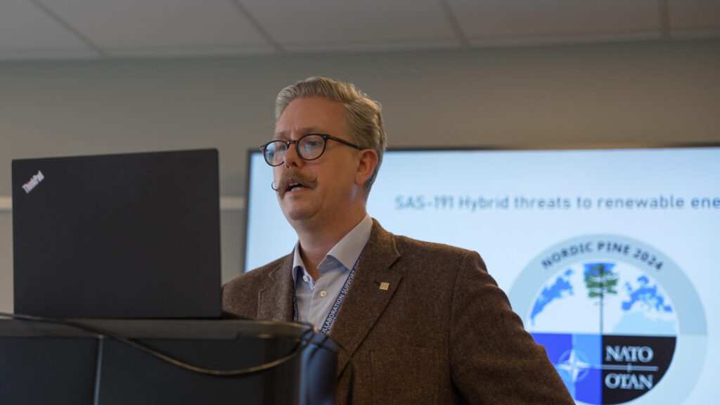 A man in a brown jacket standing in front of his laptop, speaking.