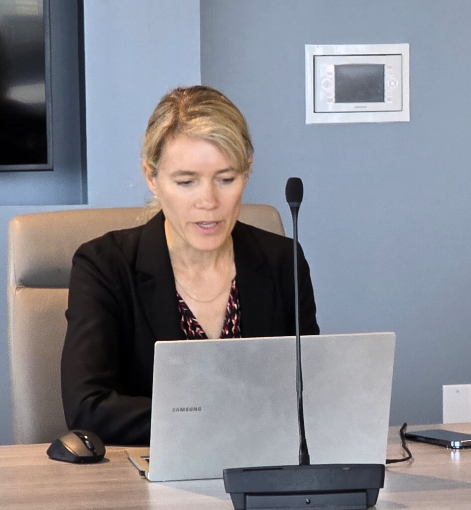 A woman in a black jacket sitting in front of her laptop, speaking.