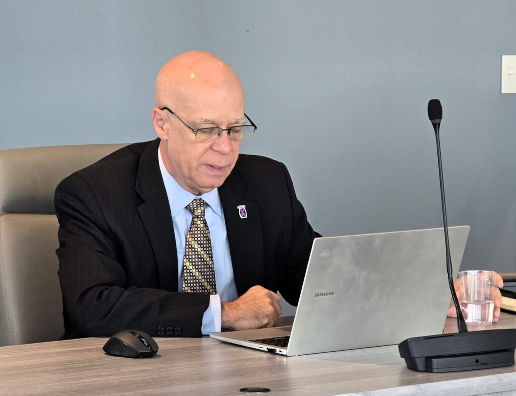 A man in a black jacket and blue shirt sitting in front of his laptop.