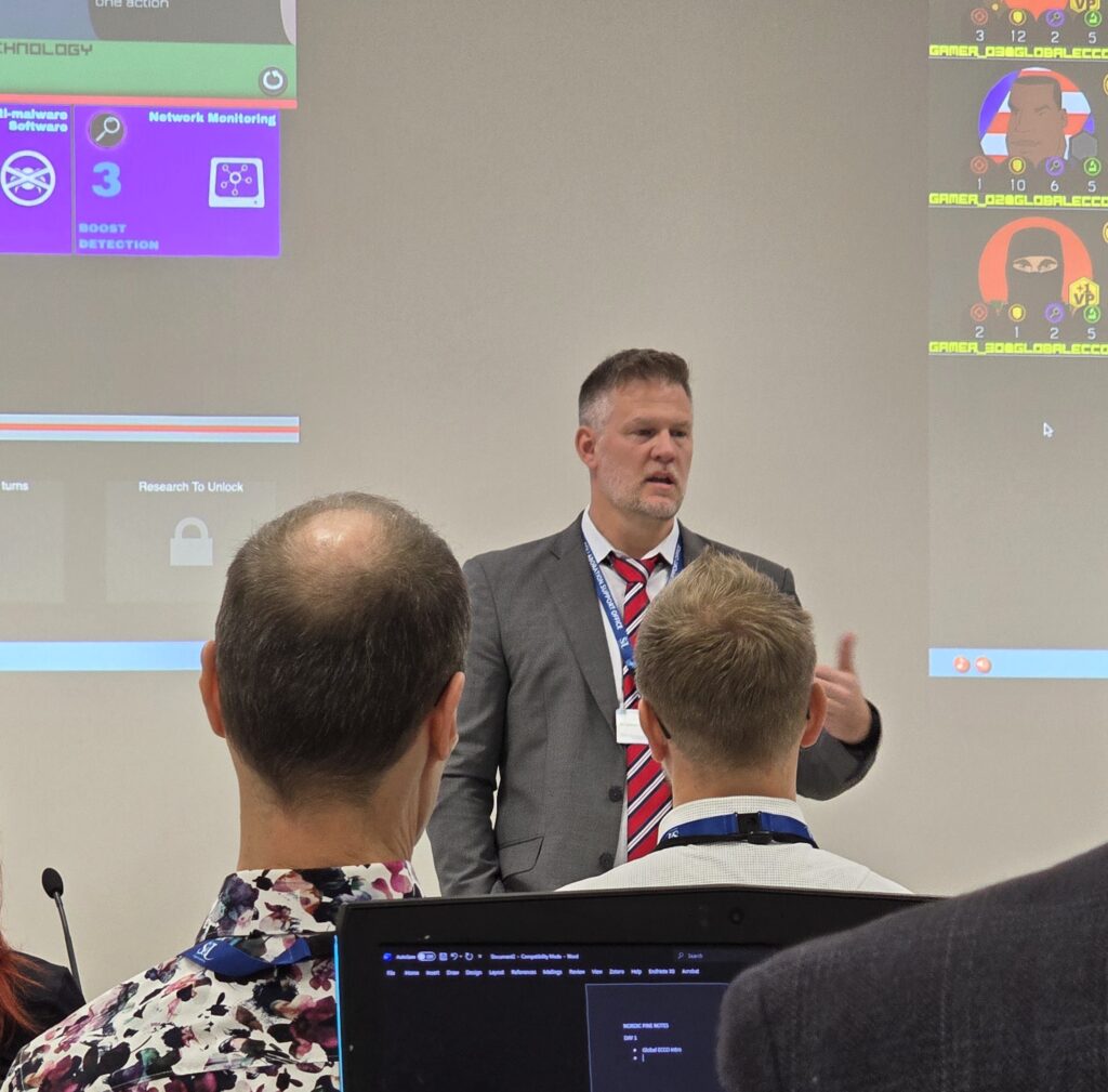 A man in a grey jacket and a red striped tie, standing in front of two people, lecturing.