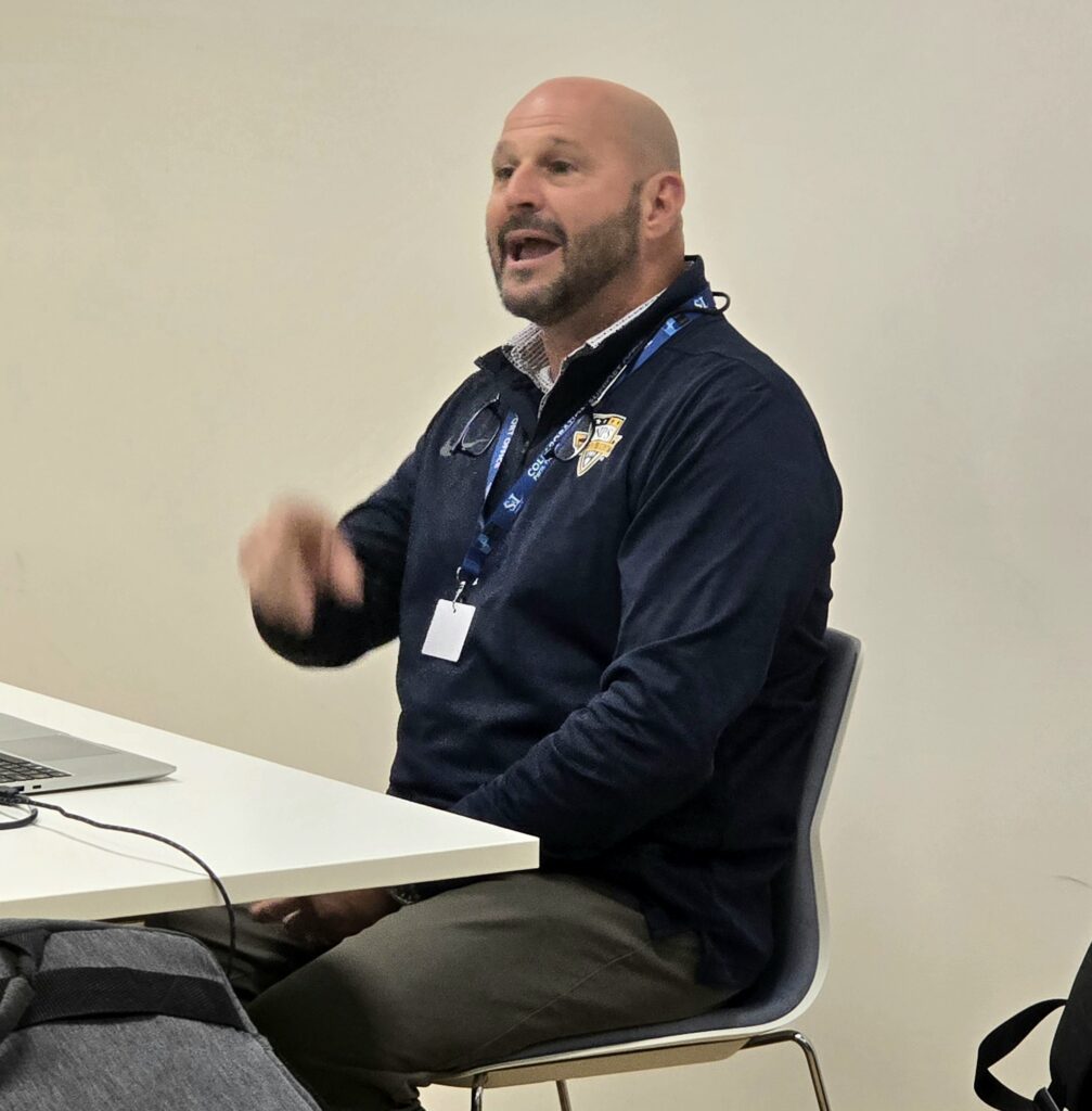 A man in a blue sweater sitting on a chair, speaking and pointing.