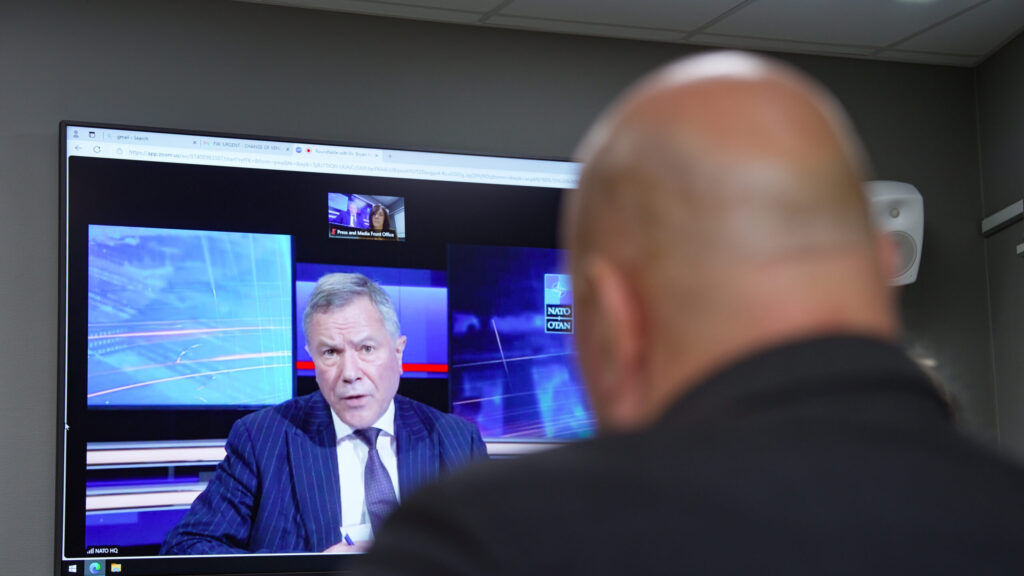 A man sitting in front of a media screen, watching another man speak.